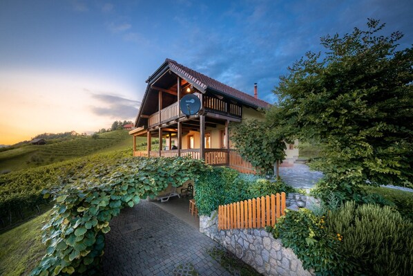 Situated on a hillside, this chalet-style building offers a balcony and a stone pathway leading to the entrance, surrounded by lush greenery and terraced vineyards.