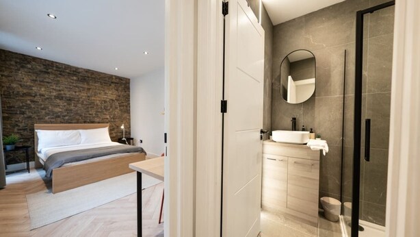 A modern bedroom with a wooden bed, a bedside table, and a stone wall. Adjacent is a bathroom with a glass shower, a sink, and a mirror. - The Battersea Scenery - Elegant 2bdr Flat (London)