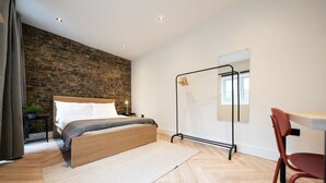 A modern bedroom with a wooden bed, a stone wall, a black metal rack, and a red chair. - The Battersea Scenery - Elegant 2bdr Flat (London)