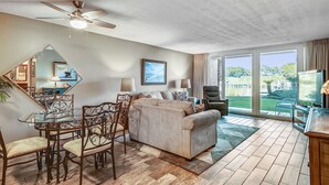 Ein geräumiger Wohnbereich mit Sofa, Esstisch und Stühlen, Deckenventilator und großen Fenstern mit Blick ins Freie.