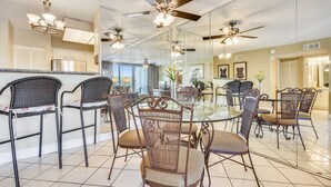 A dining area with a glass table surrounded by wicker chairs, ceiling fans, and a kitchen in the background.
