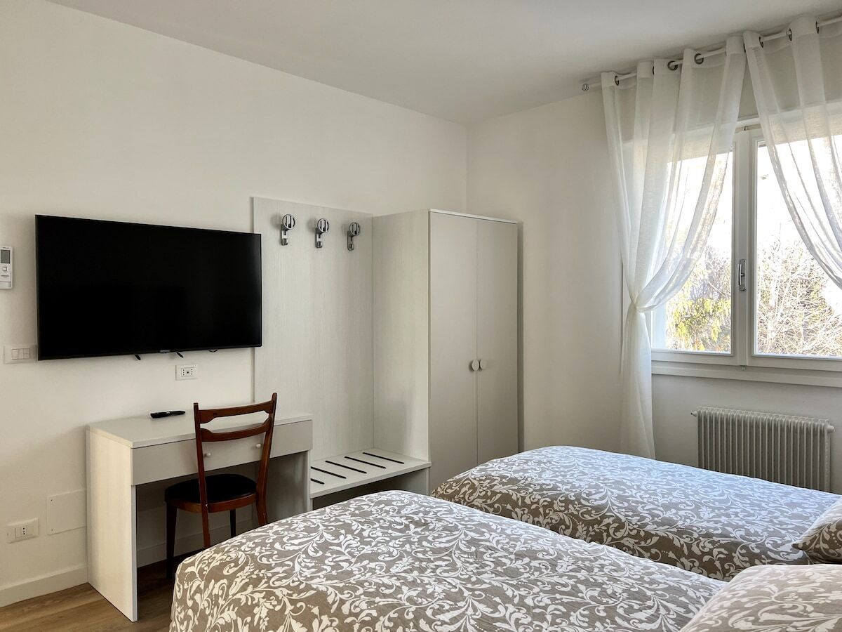 Ein Hotelzimmer mit Bett, Schreibtisch mit Stuhl, Kleiderschrank, Fernseher und einem Fenster mit Vorhängen.