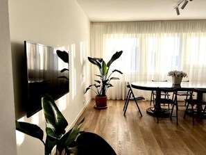 Olohuoneessa on seinälle asennettu taulutelevisio, ruokapöytä tuoleineen, ruukkukasvi ja suuri ikkuna, jossa on läpikuultavat verhot.