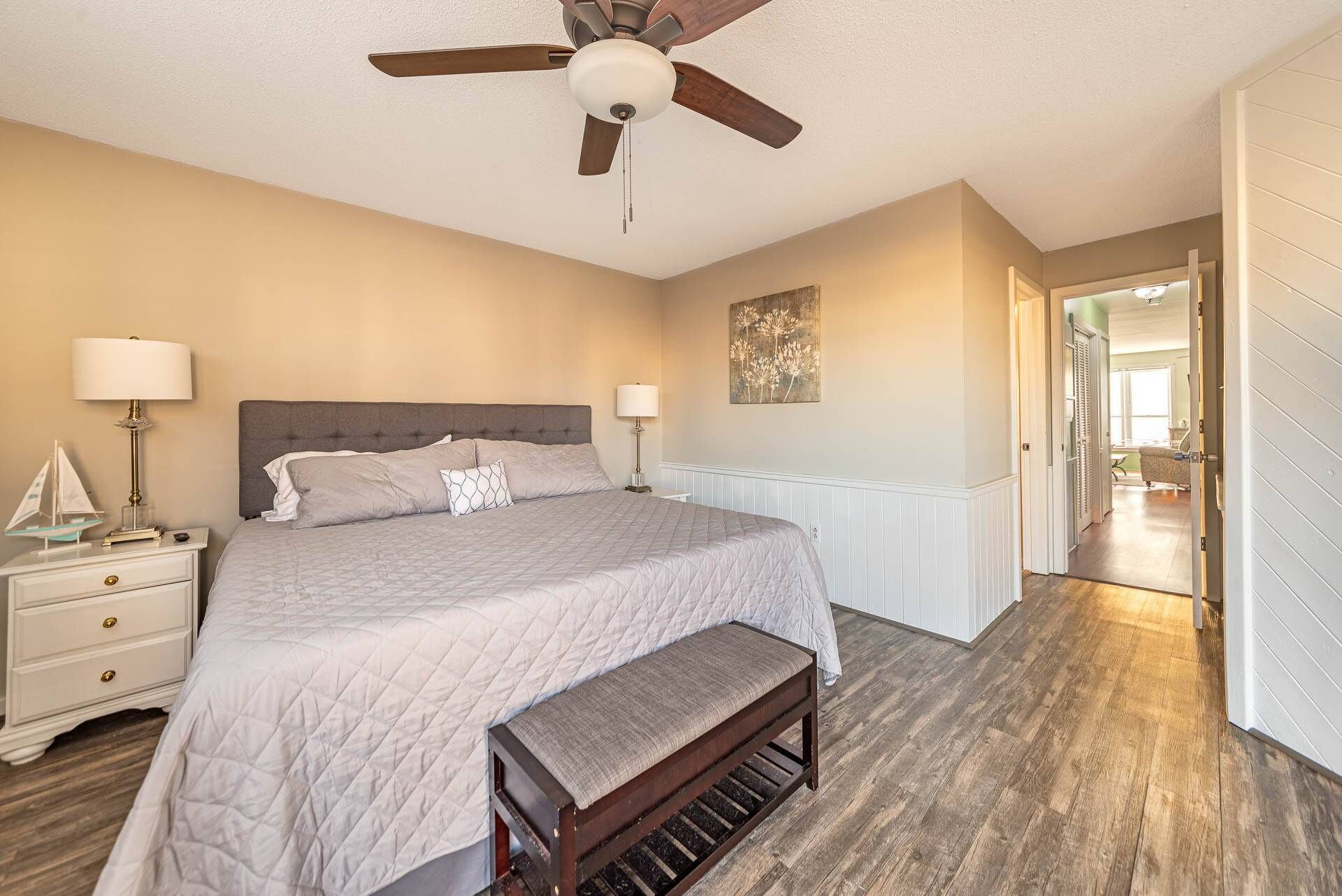 A bedroom with a bed, bedside tables, a bench, a ceiling fan, and a door leading to another room.