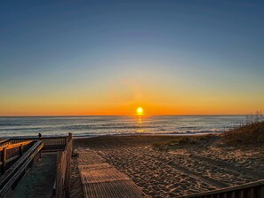 Une plage avec une promenade en bois menant à l’eau, un coucher de soleil en arrière-plan et des vagues qui viennent doucement se briser sur le rivage.