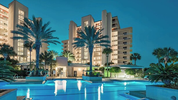A modern hotel with a pool area, palm trees, and a well-lit facade.