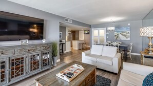 A modern living room with a flat-screen TV, a wooden cabinet, a white sofa, and a dining area with a table and chairs. - HSR 402 is a Luxury 2 BR - Completely Renovated and stunning in its decor (Destin)