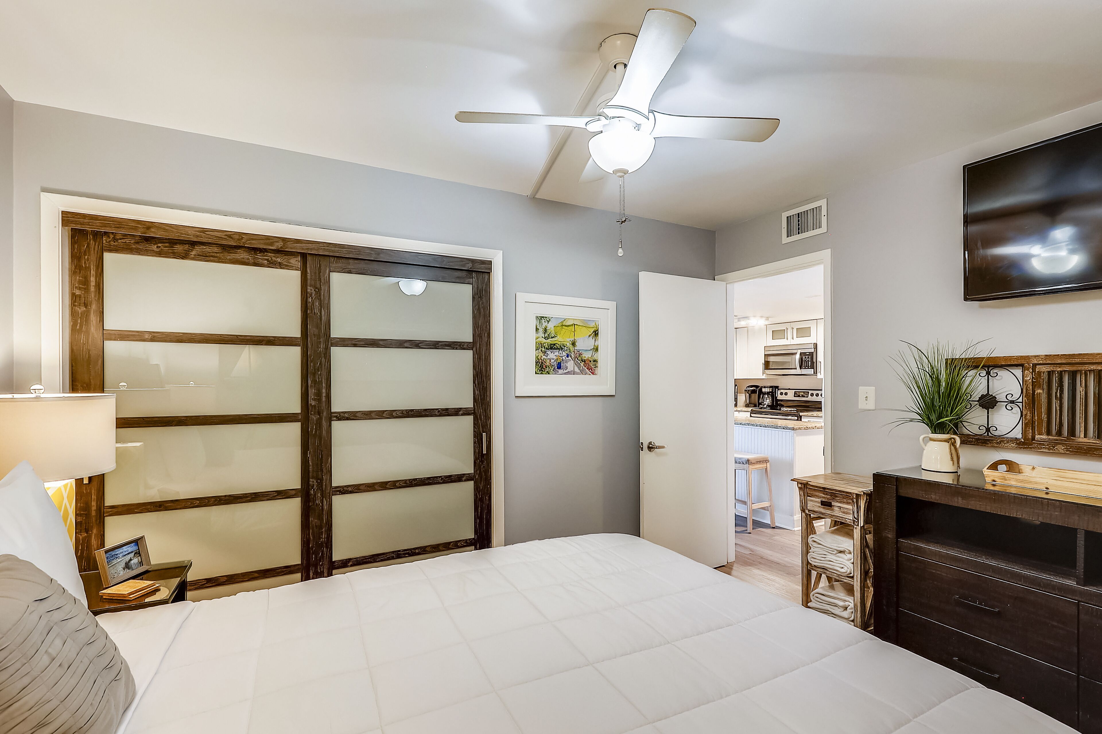 A bedroom with a bed, a ceiling fan, a wooden wardrobe, a desk with a lamp, and a kitchen visible through an open door.