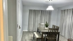 A dining area with a wooden table and chairs, a bench, a potted plant, and grey curtains. - Relax with your family in this newly renovated home away from home.  (Orange)