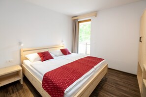 A single bed with a wooden frame, white bedding, and a red polka dot blanket. There is a small bedside table with a lamp, and a window with curtains.