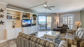 Un salon avec des canapés à rayures, un téléviseur à écran plat, un ventilateur de plafond et un balcon aménagé avec du mobilier d’extérieur.