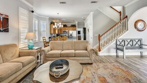 Ein geräumiges Wohnzimmer mit einem beigefarbenen Sofa, einem Couchtisch und einem Kronleuchter. Eine Treppe mit Holzwangen führt zu einer oberen Etage.