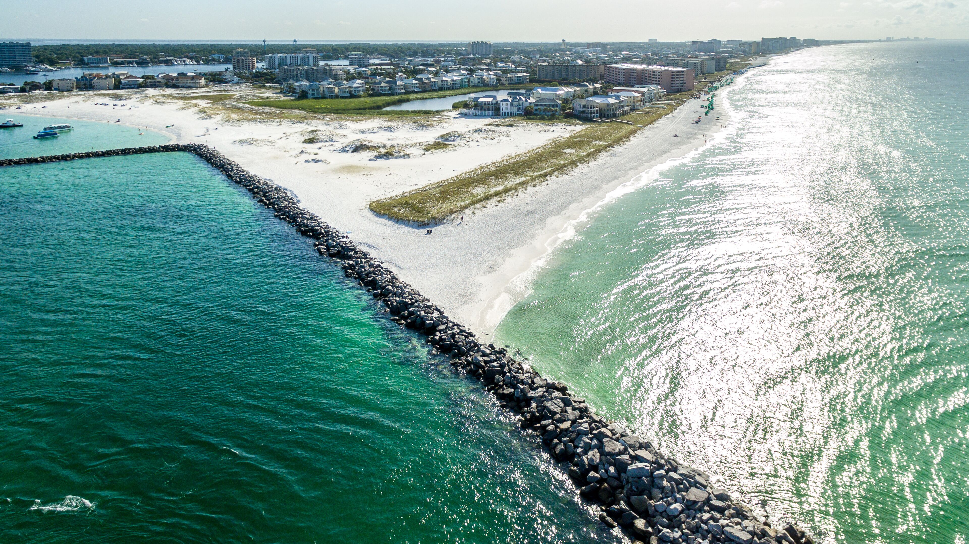 Vue aérienne d’une zone côtière avec une longue jetée s’étendant dans l’eau, une plage de sable et une rangée de bâtiments le long de la côte.