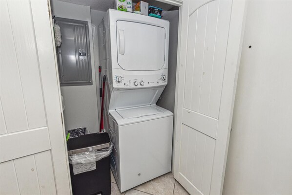 Une petite buanderie équipée d’un lave-linge et d’un sèche-linge à chargement frontal, d’une poubelle et d’un balai appuyé contre le mur.