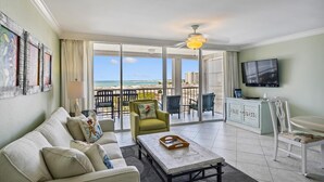Un salon spacieux comprenant un canapé, une table basse et un téléviseur. Il dispose d’un balcon avec vue sur la plage et d’un coin repas avec des chaises et un buffet.