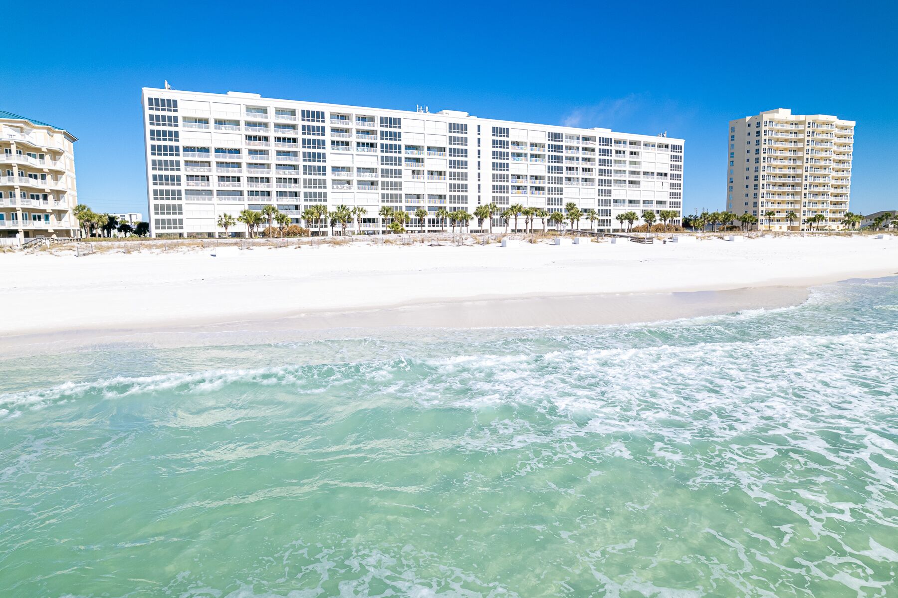 A beachfront hotel with multiple floors, a sandy beach, and palm trees.