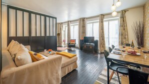 Ein Wohnzimmer mit einer Couch, einem Esstisch für zwei Personen, einem Fernseher und einem Fenster mit Vorhängen.