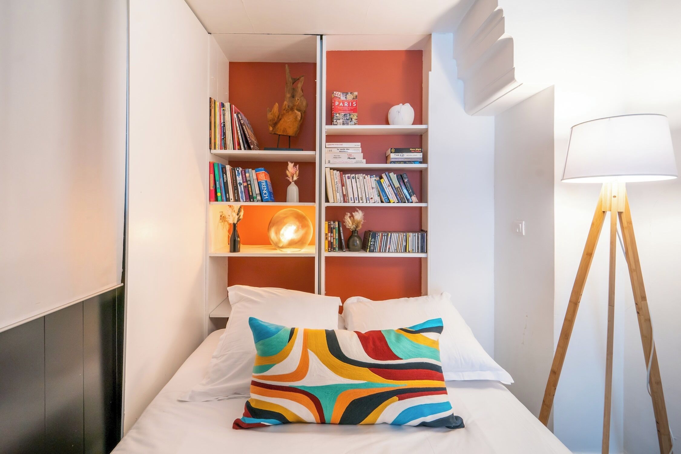A neatly arranged bedroom with a bed, colorful pillows, a bookshelf, and a floor lamp.