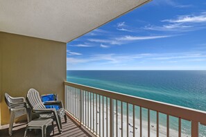 Un balcon avec deux chaises et une petite table, donnant sur une plage aux eaux d’un bleu limpide et au rivage sablonneux.