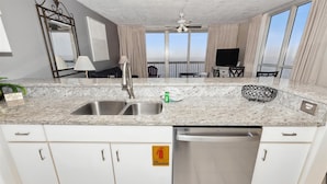 Una cocina moderna con encimera de granito, pileta de acero inoxidable y lavavajillas. Hay un cartel de extintor en la pared, y la habitación tiene ventanas grandes con cortinas.