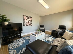 Een moderne woonkamer met een flatscreen televisie, een zwarte leren bank, een vloerkleed met patroon en een salontafel met boeken en een plant.