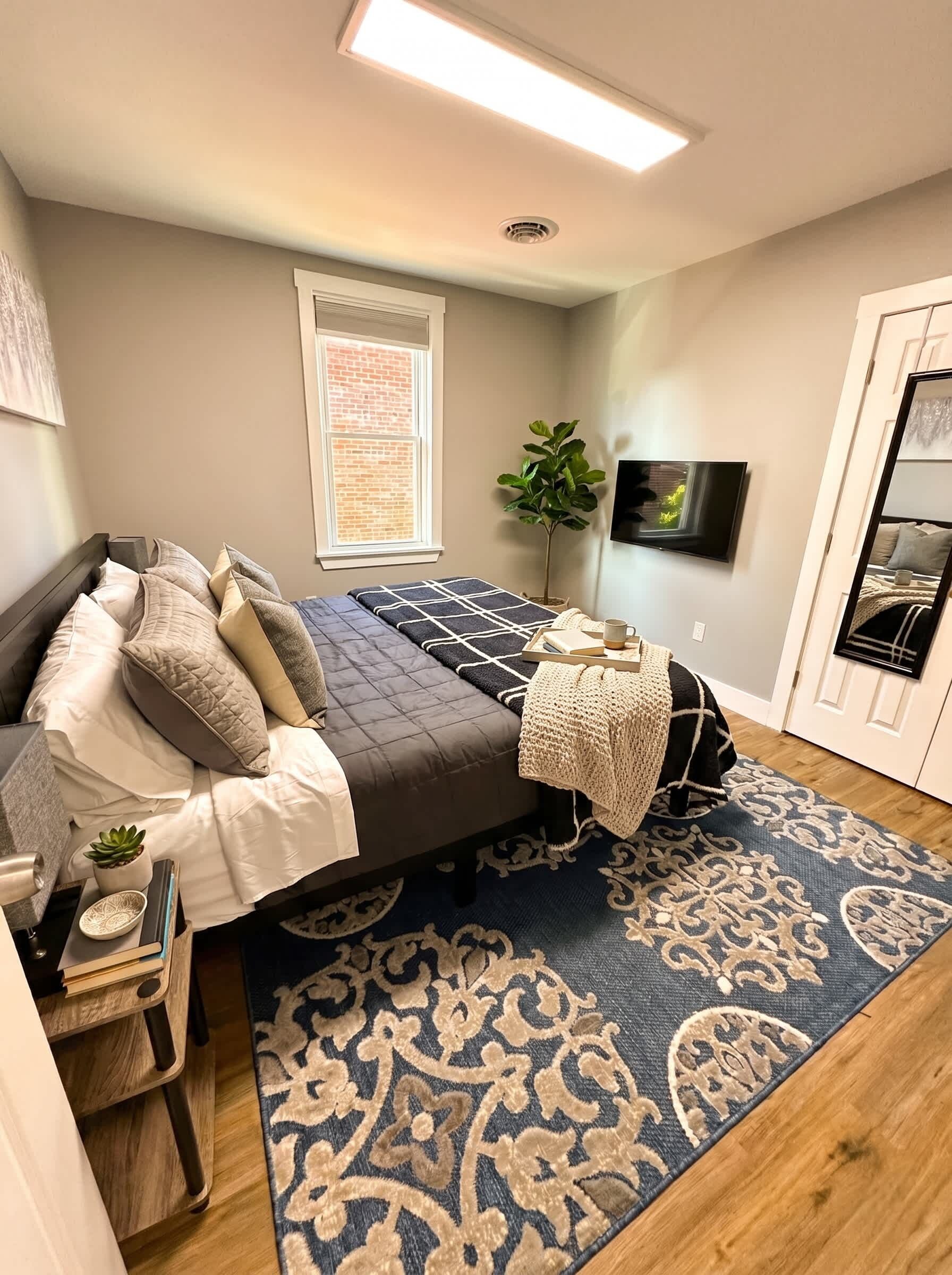 A bedroom with a bed, bedside table, lamp, potted plant, and a TV on the wall.
