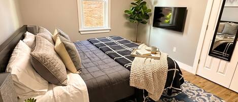 A bedroom with a bed, bedside table, lamp, potted plant, and a TV on the wall.