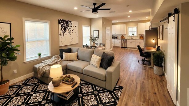 A spacious living room with a sofa, a coffee table, and a lamp. There is a kitchen in the background with white cabinets and a dining area. - Modern Loft with Skyline Views - King & Queen Beds, Close to Downtown! (Wilmington)