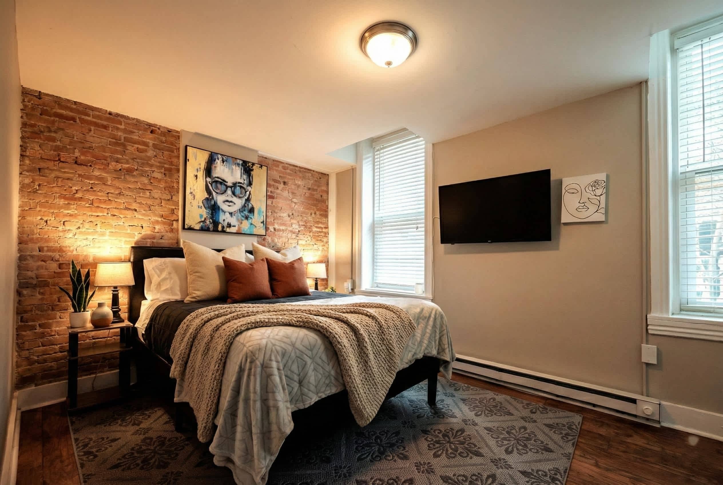 Quarto com parede de tijolos, cama com colcha estampada, mesa de cabeceira com abajur, TV fixada na parede e janela com persianas.
