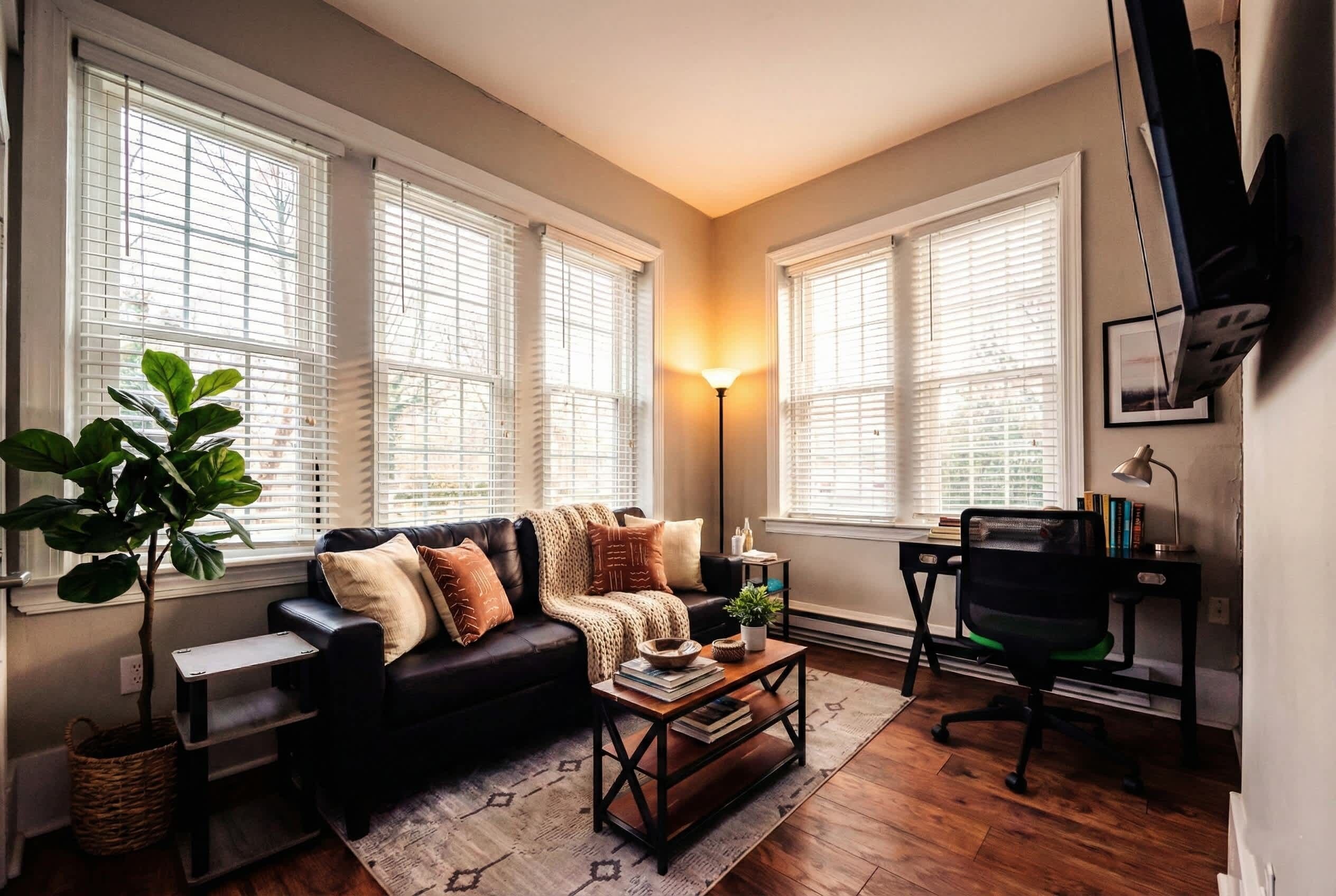 A living room with a black leather sofa, a wooden coffee table, a floor lamp, a desk with a chair, and a large window with blinds.