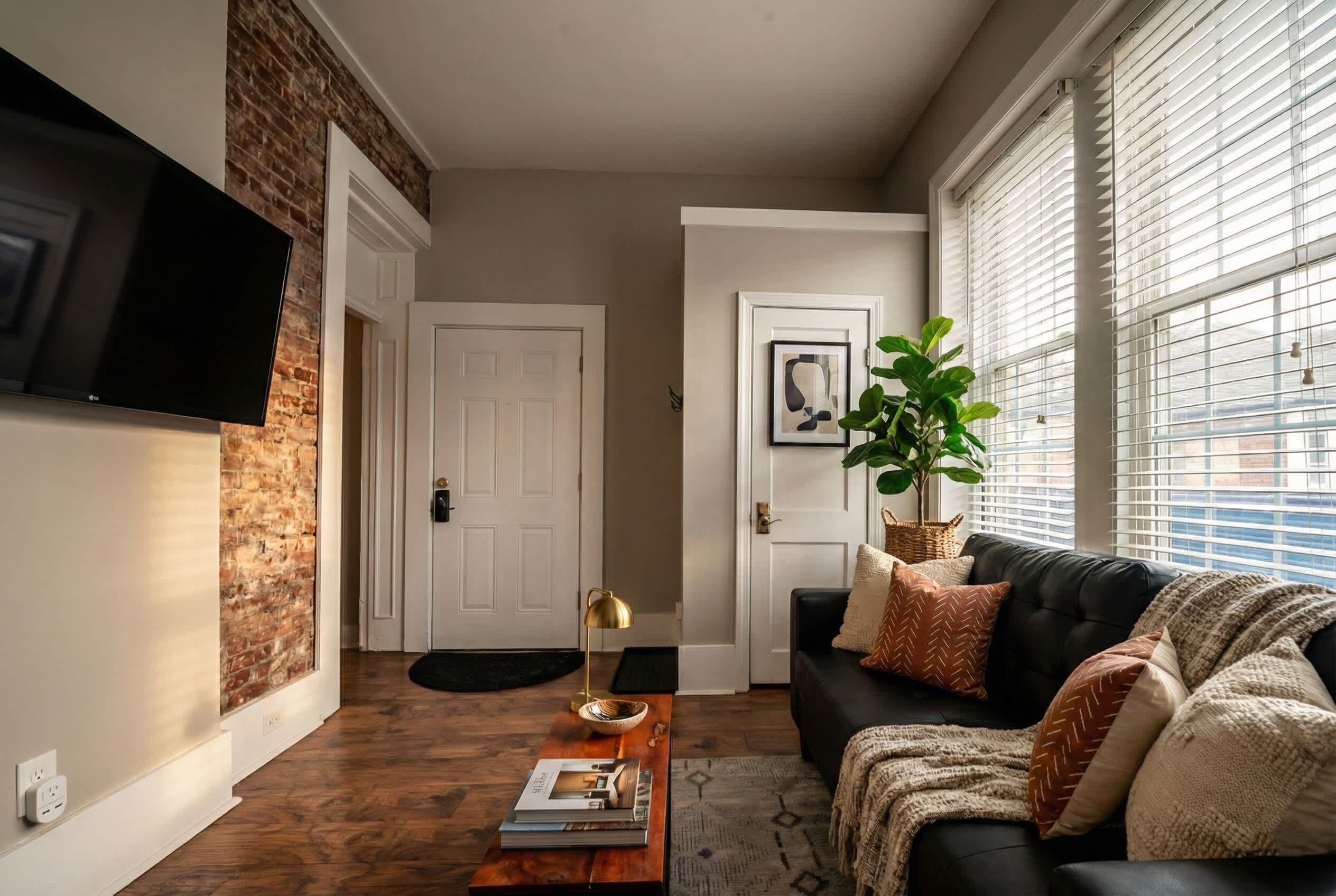 A living room with a black leather sofa, a wooden coffee table with books, a flat-screen TV mounted on a brick wall, and a large window with blinds.