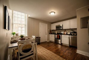 Une cuisine avec des meubles blancs, un four à micro-ondes, un four et un lave-vaisselle. Il y a une fenêtre avec des stores, une plante sur le rebord de la fenêtre et un tapis sur le sol.