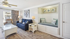Un salon avec un canapé bleu, une table basse blanche, une table d’appoint avec une lampe, un ventilateur de plafond et un banc avec des coussins.