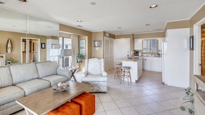 A spacious living area with a sofa, a coffee table, and a dining table. There is a kitchen area with a refrigerator, sink, and bar stools.