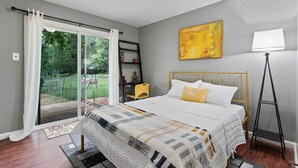 Une chambre à coucher comprenant un lit, un bureau, une chaise, une lampe sur pied et une porte-fenêtre coulissante donnant sur un jardin.