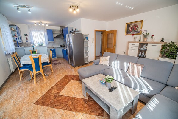 Ein modernes Wohnzimmer mit grauem Sofa, einem Esstisch mit Stühlen, einer Küche mit blauen Schränken und einem großen Fenster mit Vorhängen.
