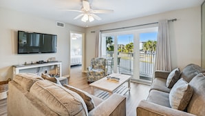 A living room with a flat-screen TV, a ceiling fan, a sofa, a coffee table, and a balcony with a view of palm trees and a parking lot. - ⛱️SeaCrest 211 - Beachfront Complex without Beachfront Prices! (Fort Walton Beach)