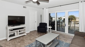 A living room with a flat-screen TV, a dark armchair, a small white table, and a balcony with a view of palm trees and buildings.
