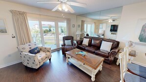 A spacious living room with a brown leather sofa, a patterned armchair, and a glass coffee table. There is a ceiling fan, a large window with curtains, and a view of the outside.