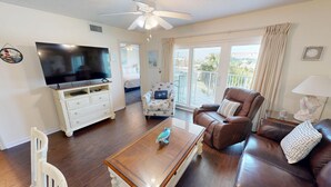 A living room with a TV, a wooden coffee table, a brown leather sofa, a white armchair, and a balcony with curtains.