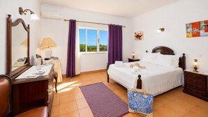 A bedroom with a bed, a desk, a chair, a window with curtains, a wall-mounted air conditioner, and a painting. - Villa Sol Nascente - Four Bedroom Villa, Sleeps 8 (Porches)