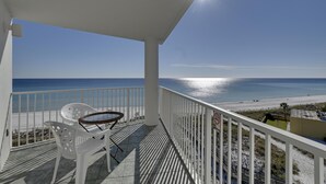 Uma varanda com vista para a praia, cadeiras brancas e uma mesa pequena.