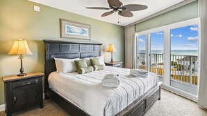 Une chambre d’hôtel avec un grand lit, deux tables de chevet avec des lampes, un ventilateur de plafond, une vue sur l’océan par de grandes fenêtres et un tableau encadré au mur.