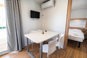 Une pièce de taille modeste, avec une table blanche, deux chaises blanches, un téléviseur à écran plat fixé au mur, un lit superposé en bois et une fenêtre ornée d’un rideau à motifs noir et blanc.