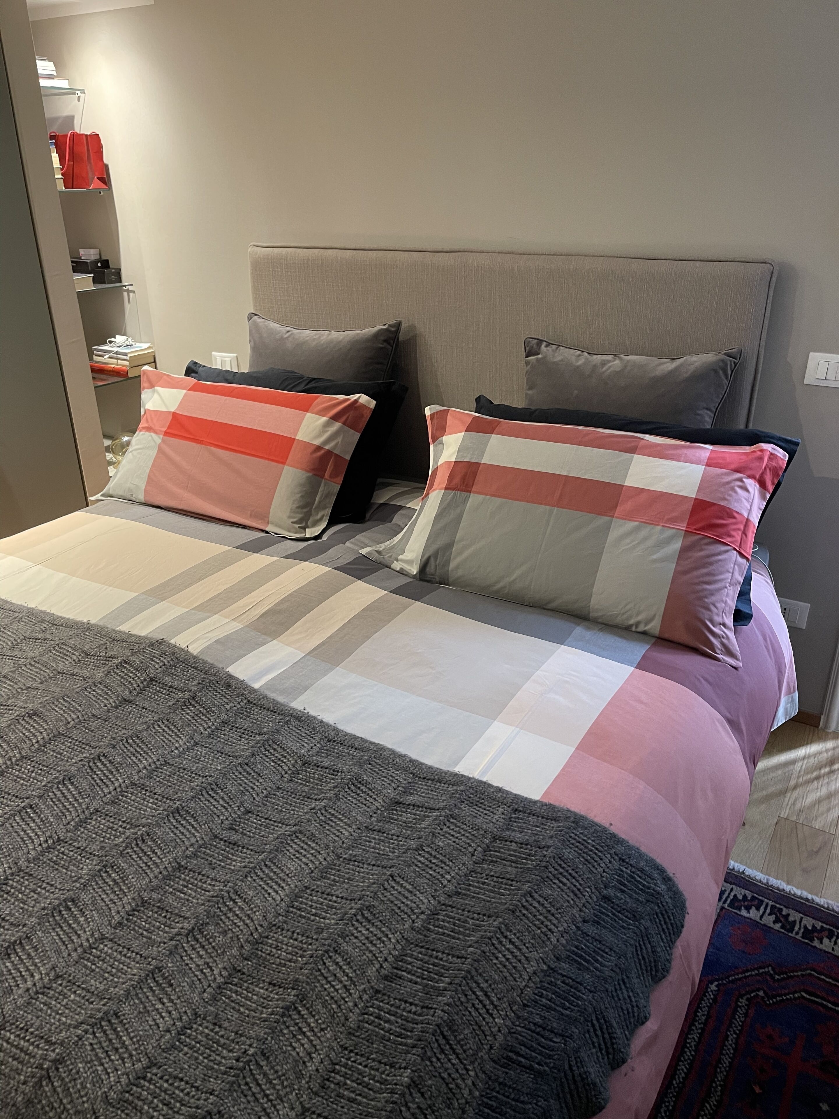 Ein ordentlich gemachtes Bett mit einer karierten Tagesdecke, zwei Kissen, einem grauen Kopfteil und einer grauen Strickdecke.