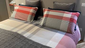 Ein ordentlich gemachtes Bett mit einer karierten Tagesdecke, zwei Kissen, einem grauen Kopfteil und einer grauen Strickdecke.