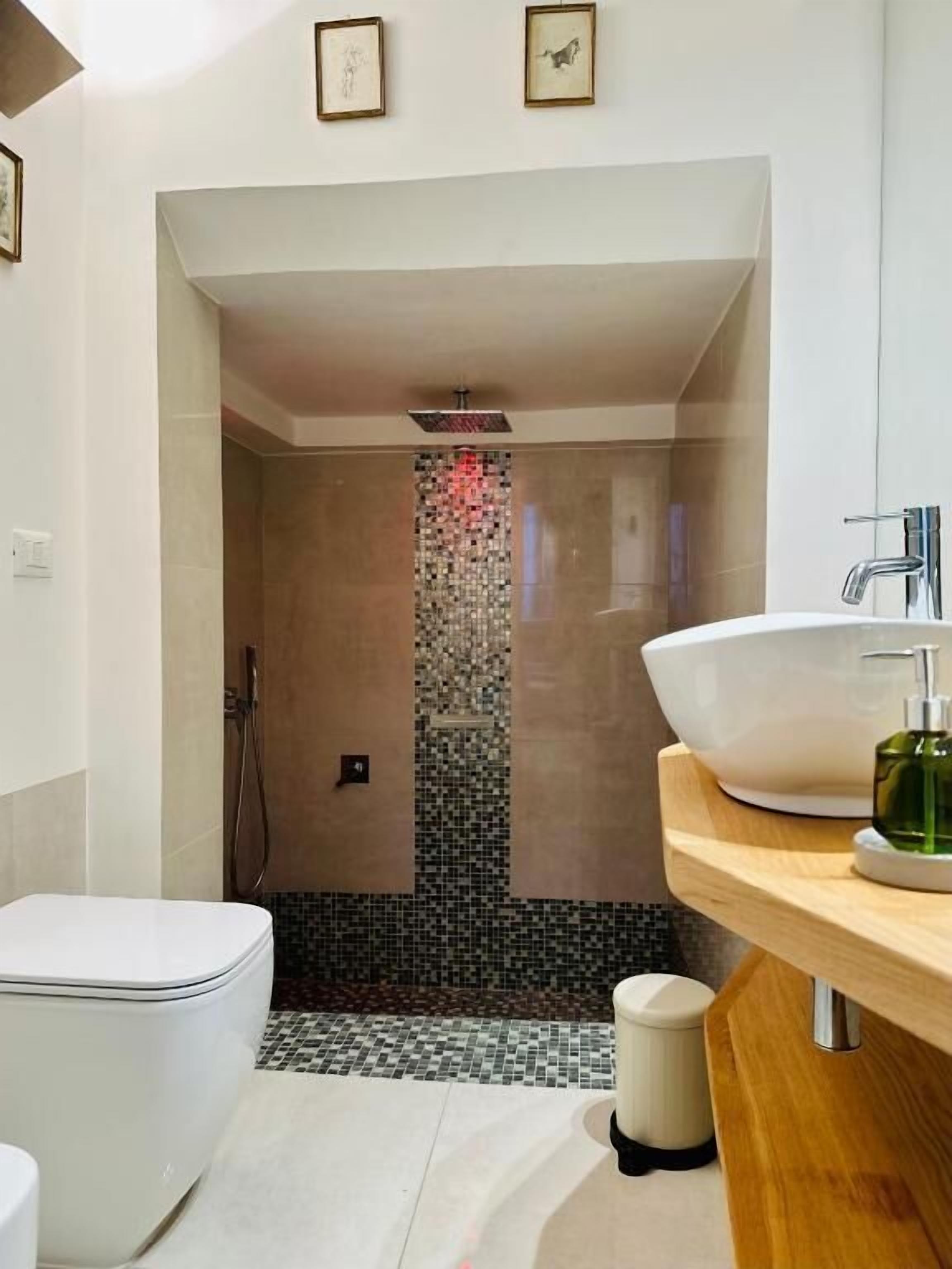 Ein modernes Badezimmer mit Toilette in Weiß, einem großen Waschbecken mit grünem Seifenspender und einer Duschkabine mit Wand aus Mosaikfliesen.