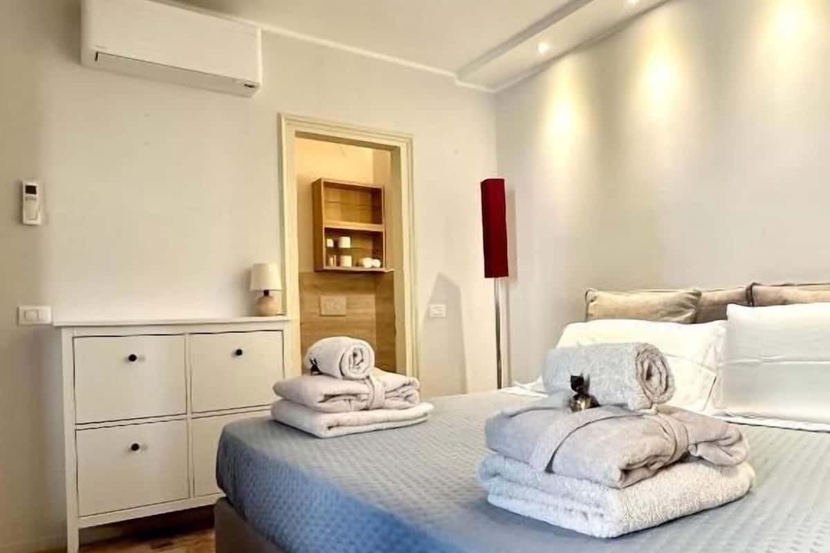 Ein Schlafzimmer mit Bett, Nachttisch, Lampe, Kleiderschrank und einem Regal mit Gegenständen.