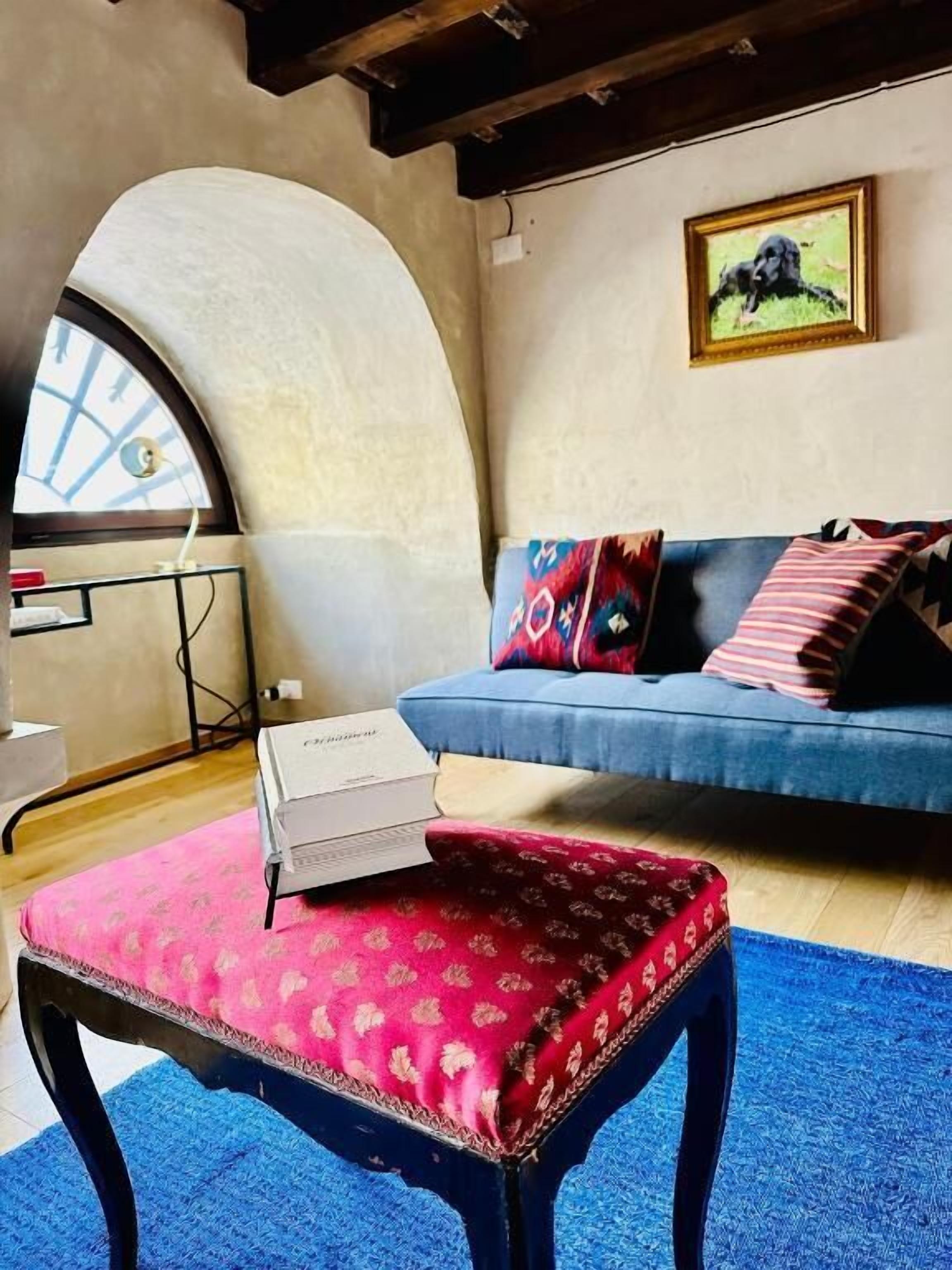 Ein Zimmer mit einem blauen Sofa, einem rot gemusterten Hocker, einem gerahmten Bild an der Wand und einem Oberlicht.