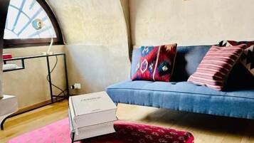 Ein Zimmer mit einem blauen Sofa, einem rot gemusterten Hocker, einem gerahmten Bild an der Wand und einem Oberlicht.
